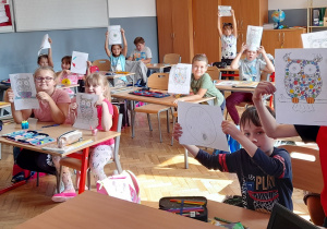 Uczniowie klasy 2b siedzą w ławkach i z uśmiechami na twarzy pokazują wykonane przez siebie prace: kolorowe duże kropki lub kolorowe sowy z stworzone z małych kropek.
